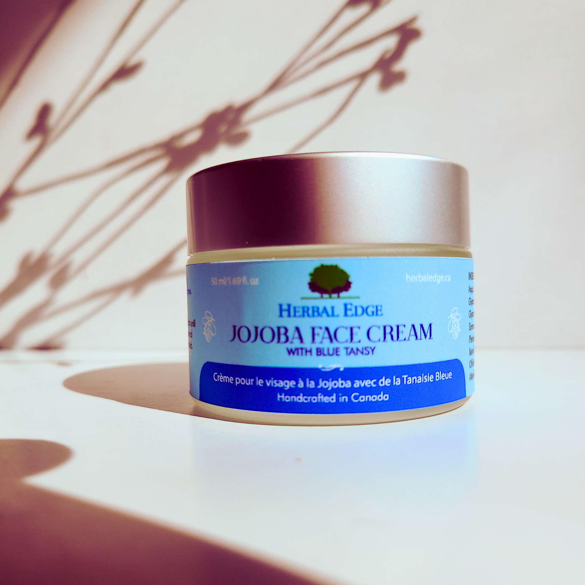 A jar of Herbal Edge Jojoba Face Cream with Blue Tansy placed on a white surface with a shadow of a plant leaf on it. The label on the jar indicates that the product is handcrafted in Canada.