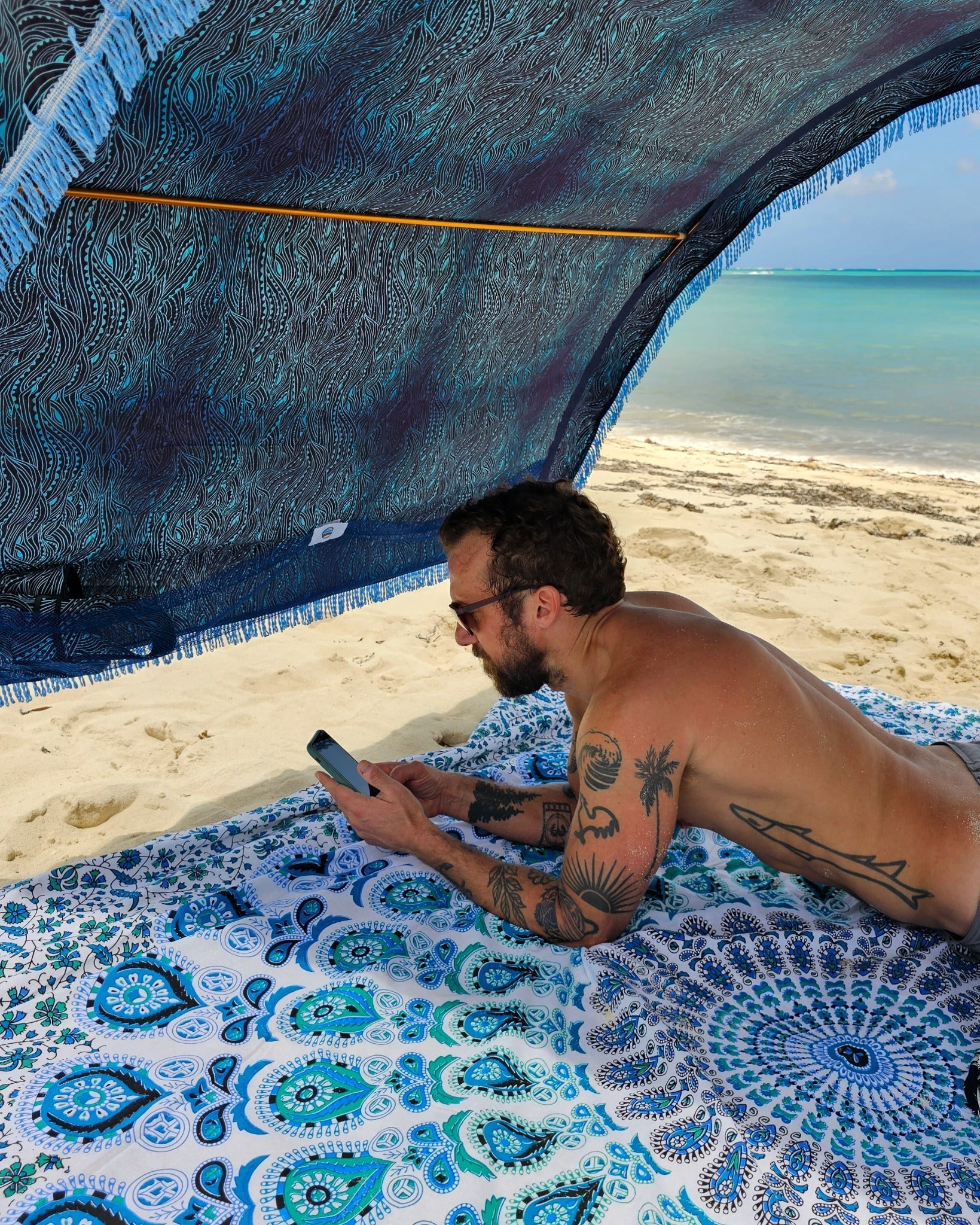 Man using his phone beneath a Suniela portable beach cabana with Ocean Waves cotton canopy on a sandy beach