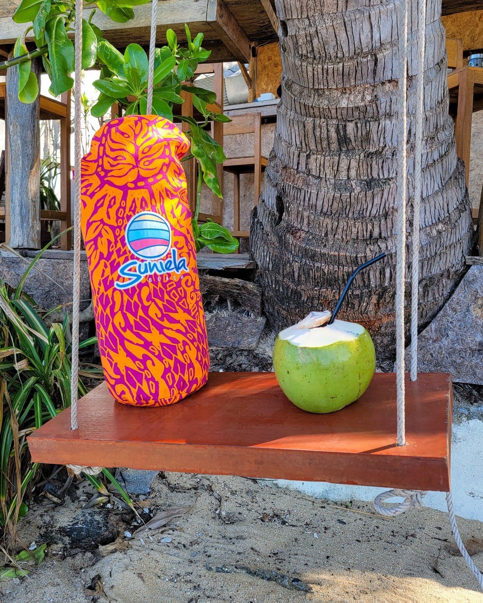 Suniela Tribal Jungle cabana packed in its carry bag resting on a wooden swing beside a fresh coconut drink, highlighting the compact size and portability of the beach cabana