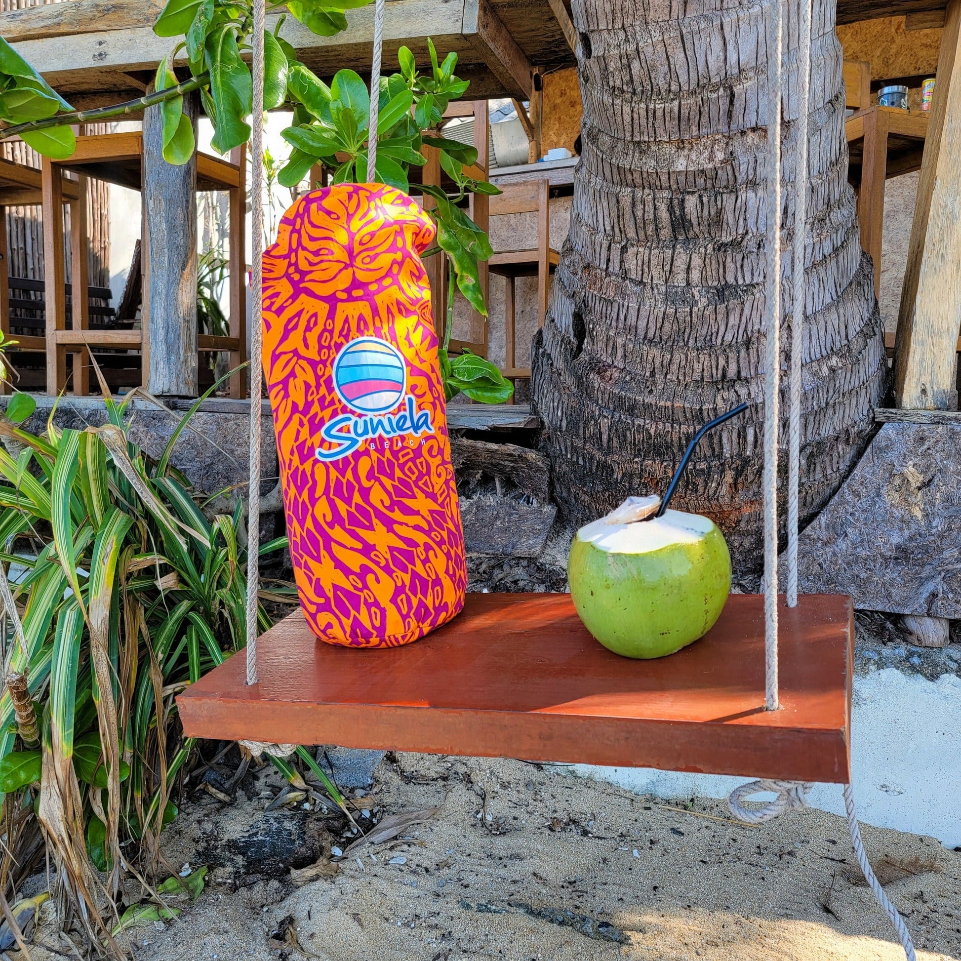 Suniela Tribal Jungle cabana packed in its carry bag resting on a wooden swing beside a fresh coconut drink, highlighting the compact size and portability of the beach cabana