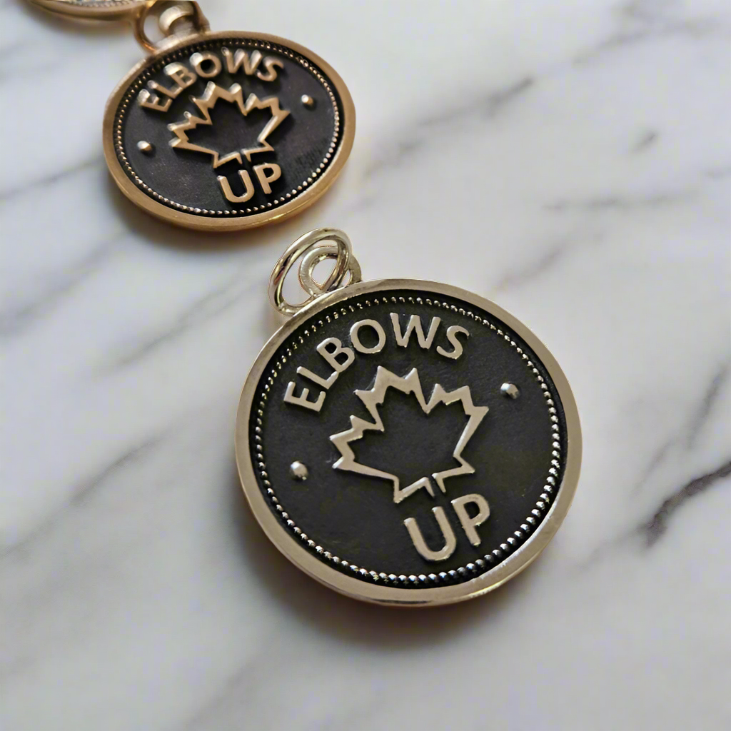 Two coin pendants with 'Elbows Up' and a maple leaf design, one in gold and the other in silver, placed on a marble surface.