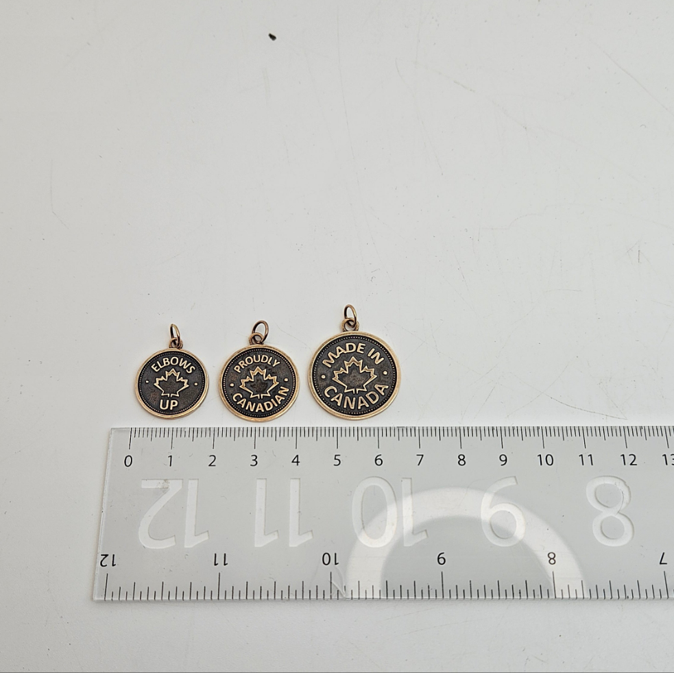Three coin pendants, of increasing sizes, with 'Elbows Up' or 'Proudly Canadian' or 'Made in Canada' and a maple leaf design, in gold above a ruler on white background