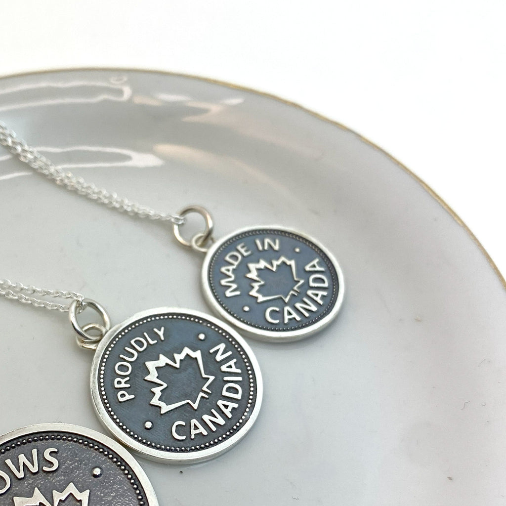 Sterling silver coin pendants of different sizes with a 'ELBOWS UP', 'PROUDLY CANADIAN', or 'MADE IN CANADA' inscription and a maple leaf design, displayed on a white plate.