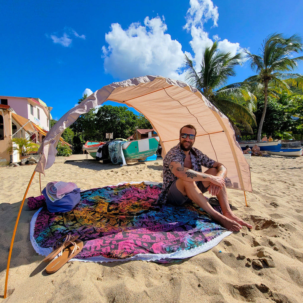 A man sits on a colorful patterned blanket under a beige beach canopy on a sandy beach. A blue bag, sandals, palm trees, boats, and a bright blue sky are visible in the background.