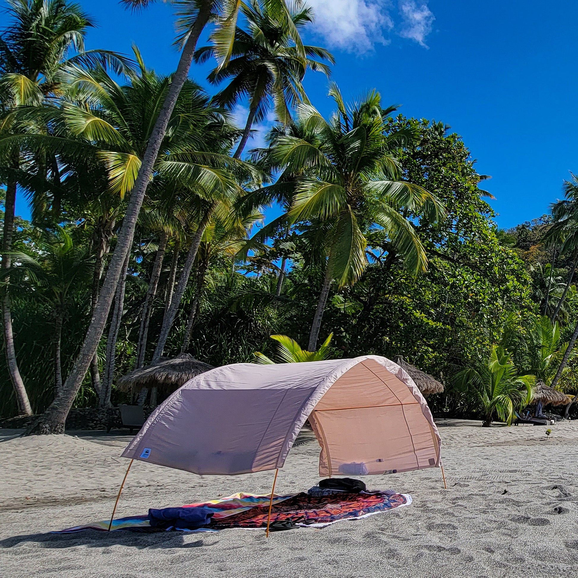 An arched cabana tent with a sand coloured canopy is pitched on a sandy beach under a bright blue sky with tall palm trees in the background.