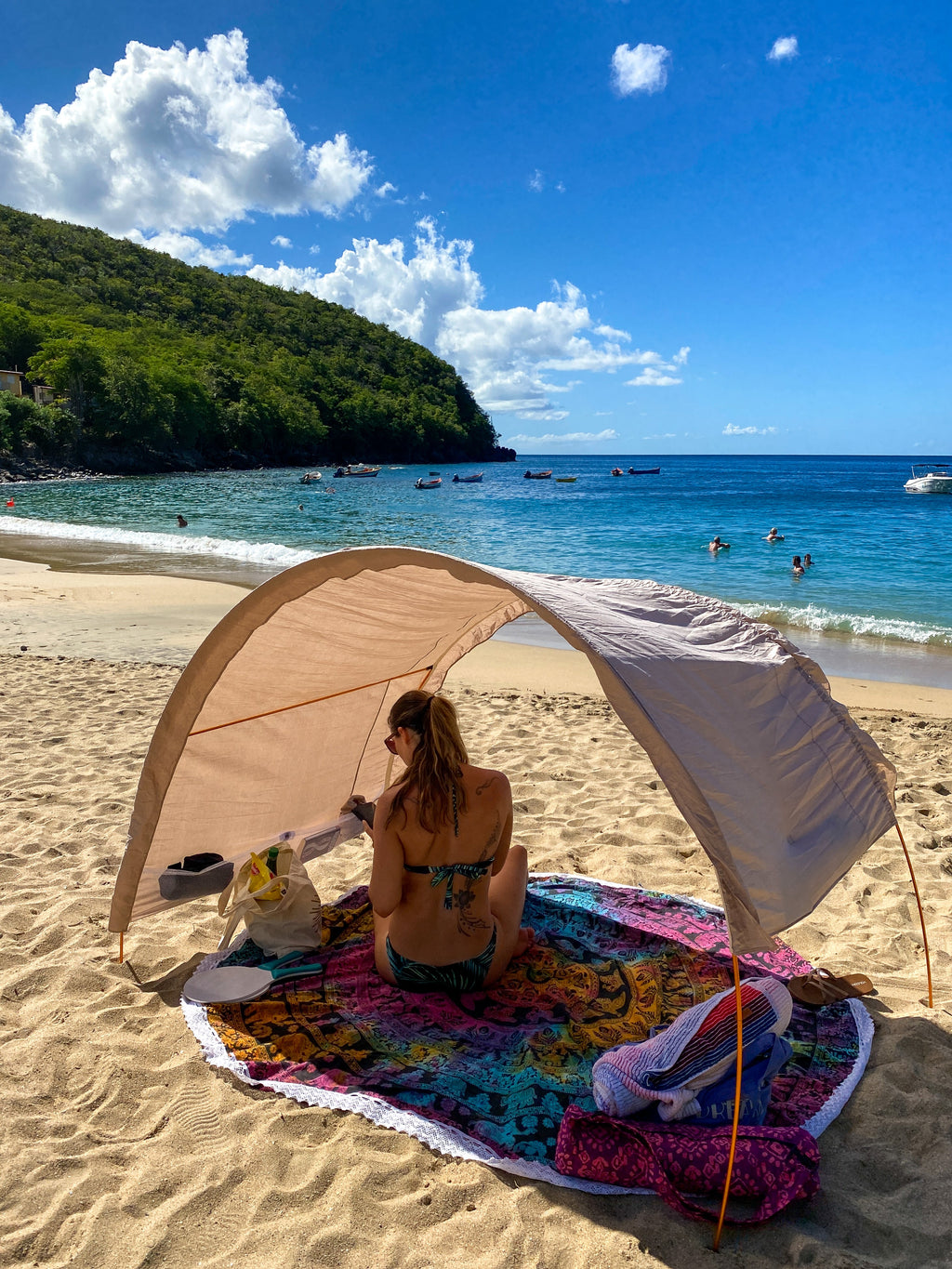 A woman sits under a beige Suniela  beach canopy on a colorful patterned blanket on a sandy beach, with a tropical ocean, green hill, and blue sky in the background.