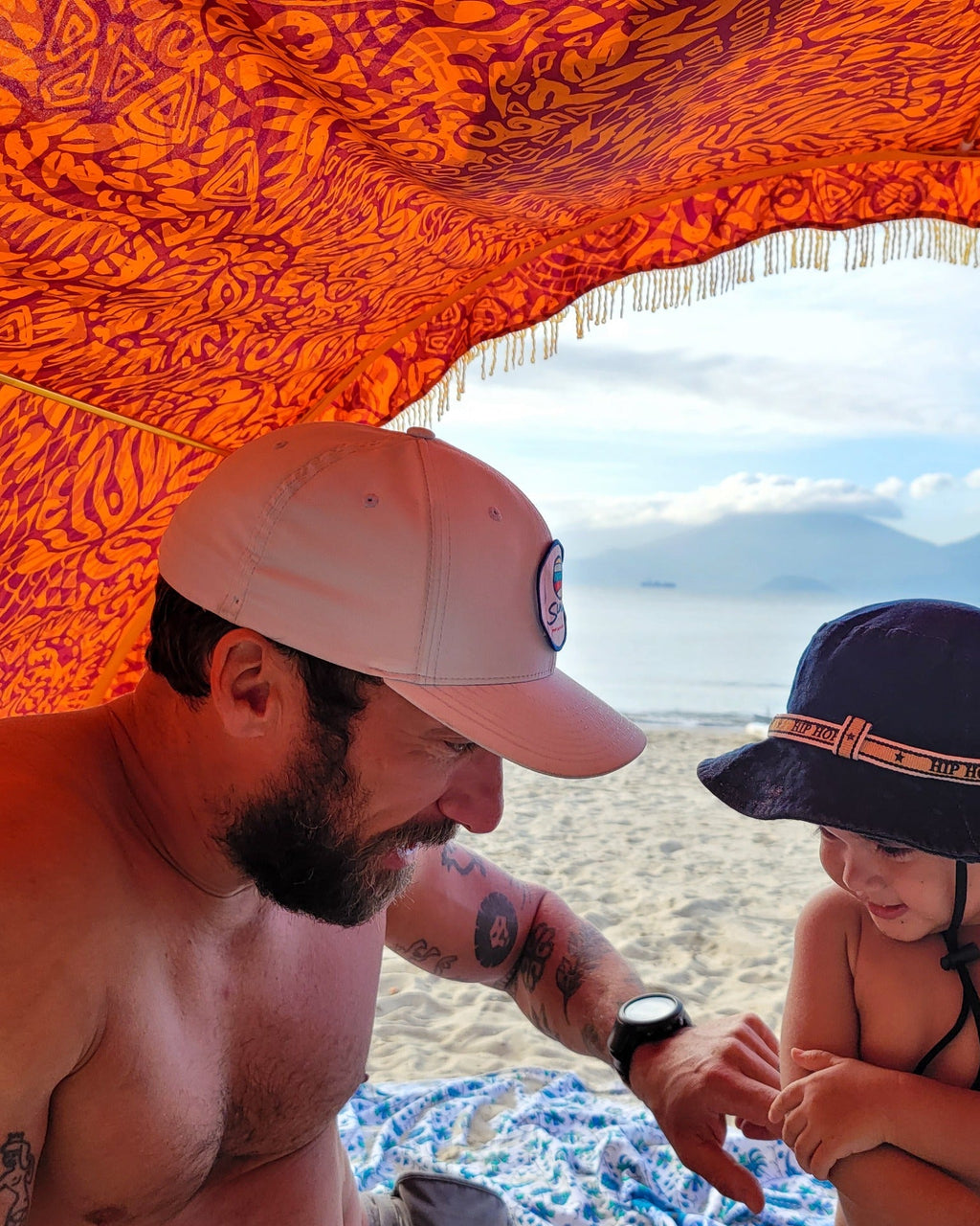 Adult and child sitting beneath a Suniela Tribal Jungle cabana on the beach, sheltered in the shade, with the ocean in the background