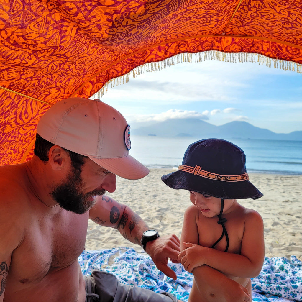Adult and child sitting beneath a Suniela Tribal Jungle cabana on the beach, sheltered in the shade, with the ocean in the background