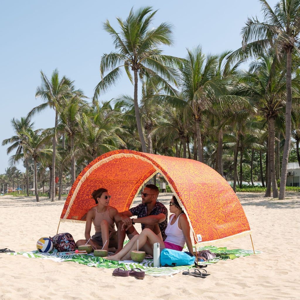Friends relaxing under a Suniela beach cabana with the Tribal Jungle orange cotton canopy and fringe on a palm-lined beach