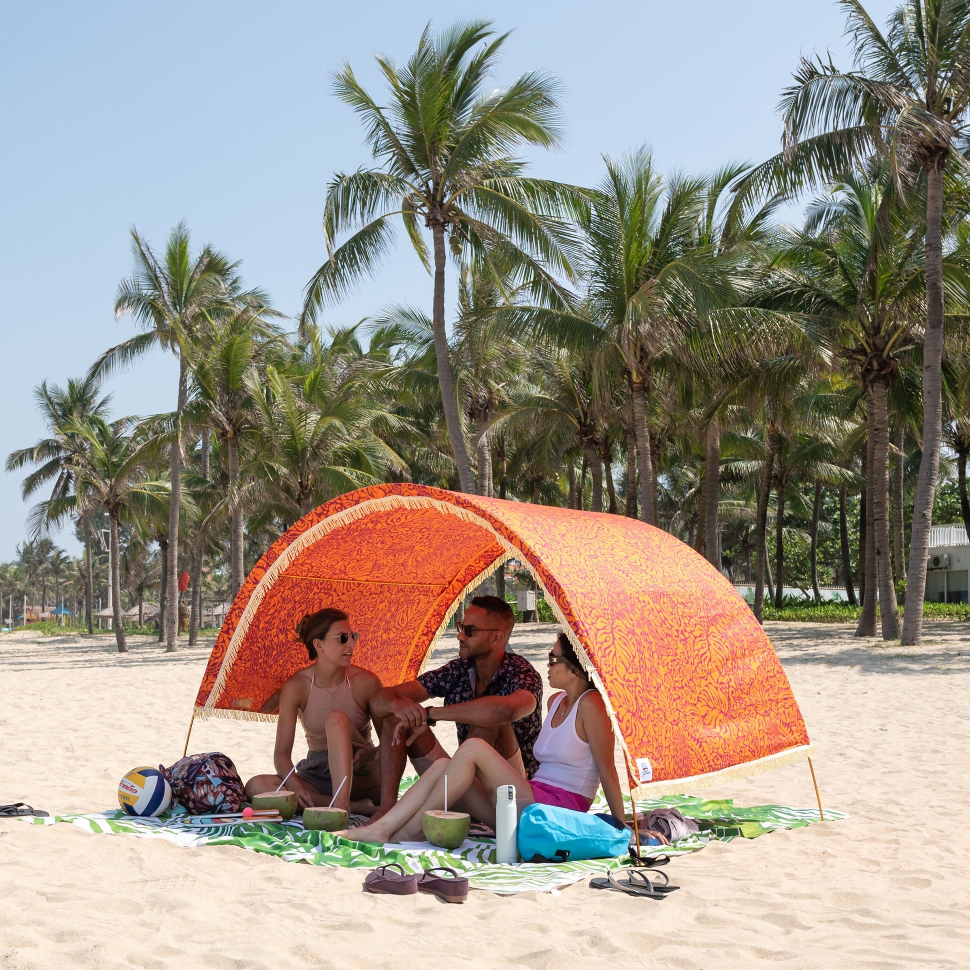 Friends relaxing under a Suniela beach cabana with the Tribal Jungle orange cotton canopy and fringe on a palm-lined beach