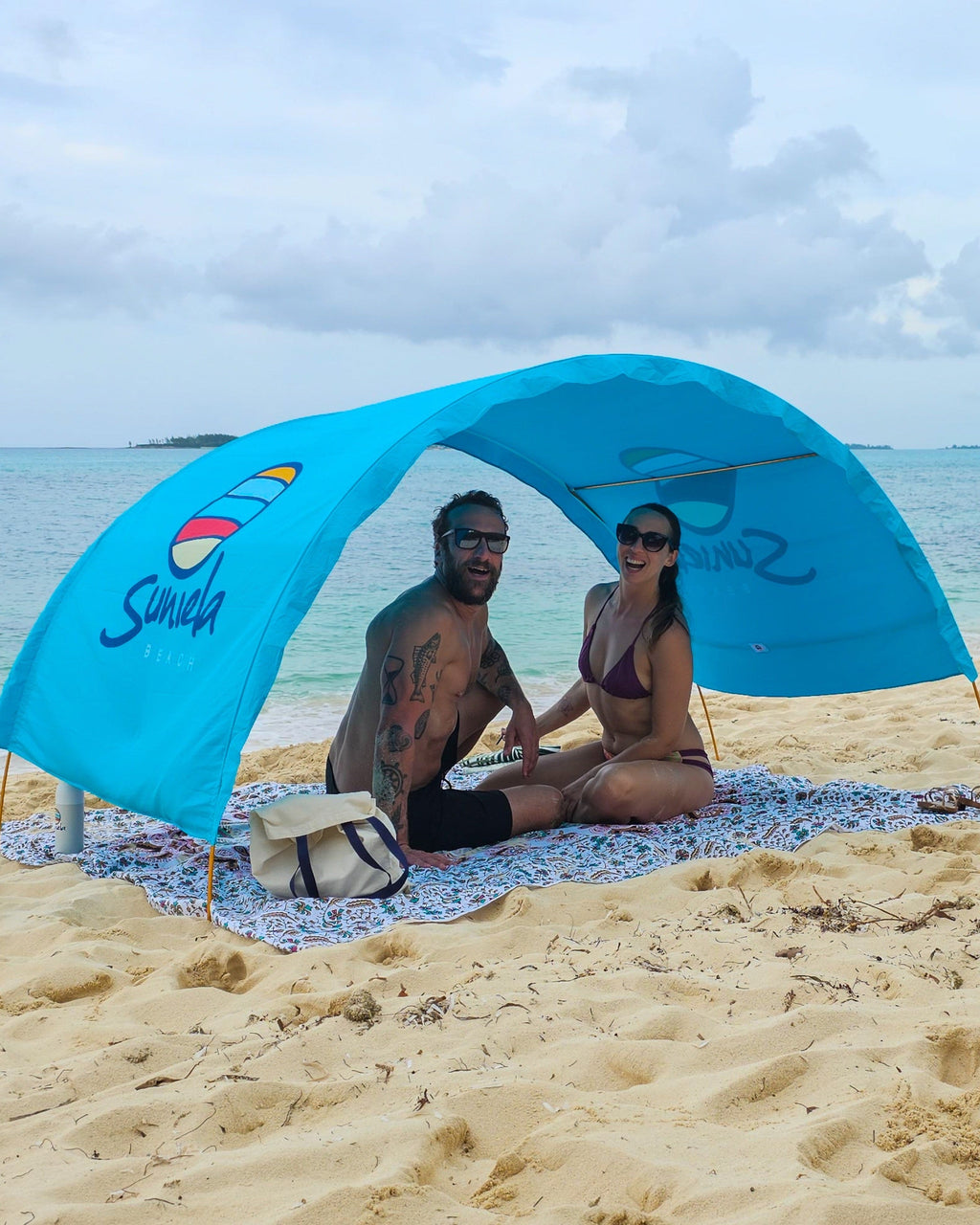 Couple sitting beneath an arched, blue Suniela shade cabana on a sandy beach