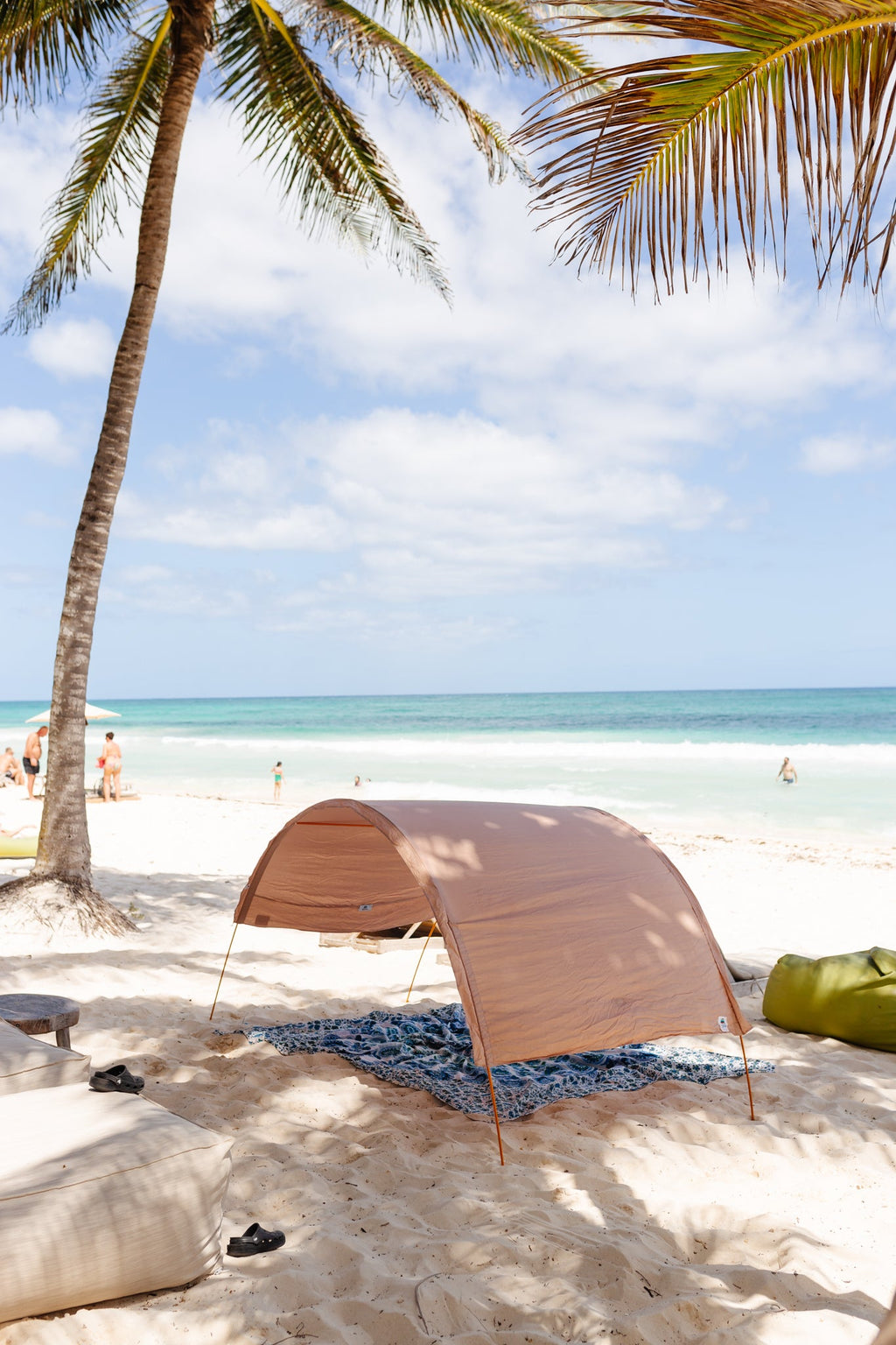 A tan beach canopy tent set up on white sand at a tropical beach, with a palm tree, ocean, and sky in the background.