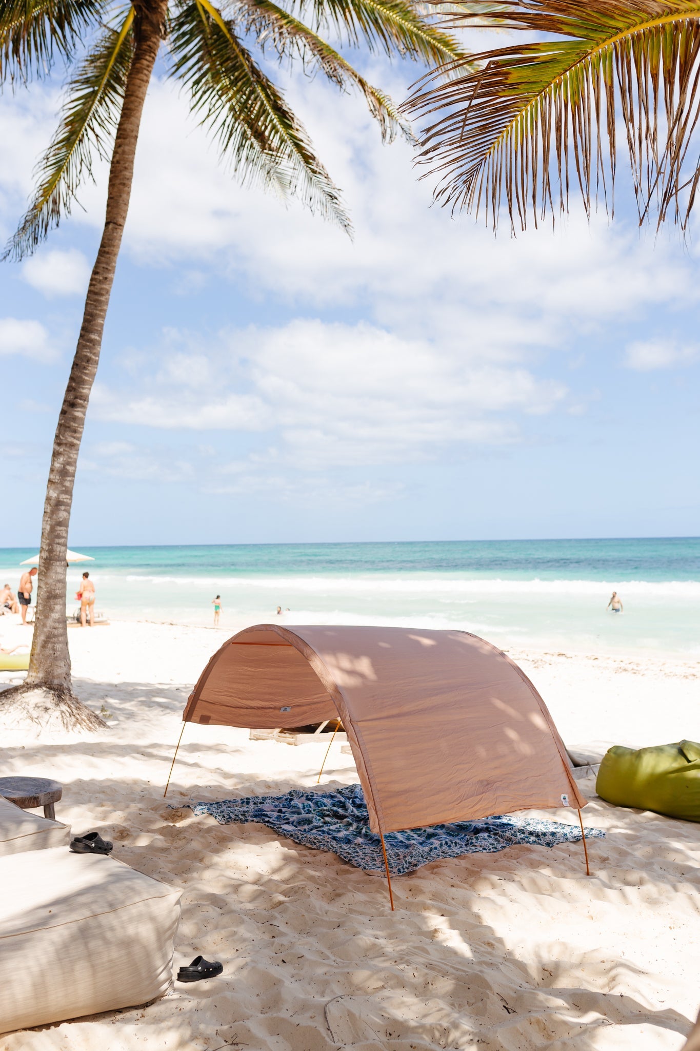 A tan beach canopy tent set up on white sand at a tropical beach, with a palm tree, ocean, and sky in the background.