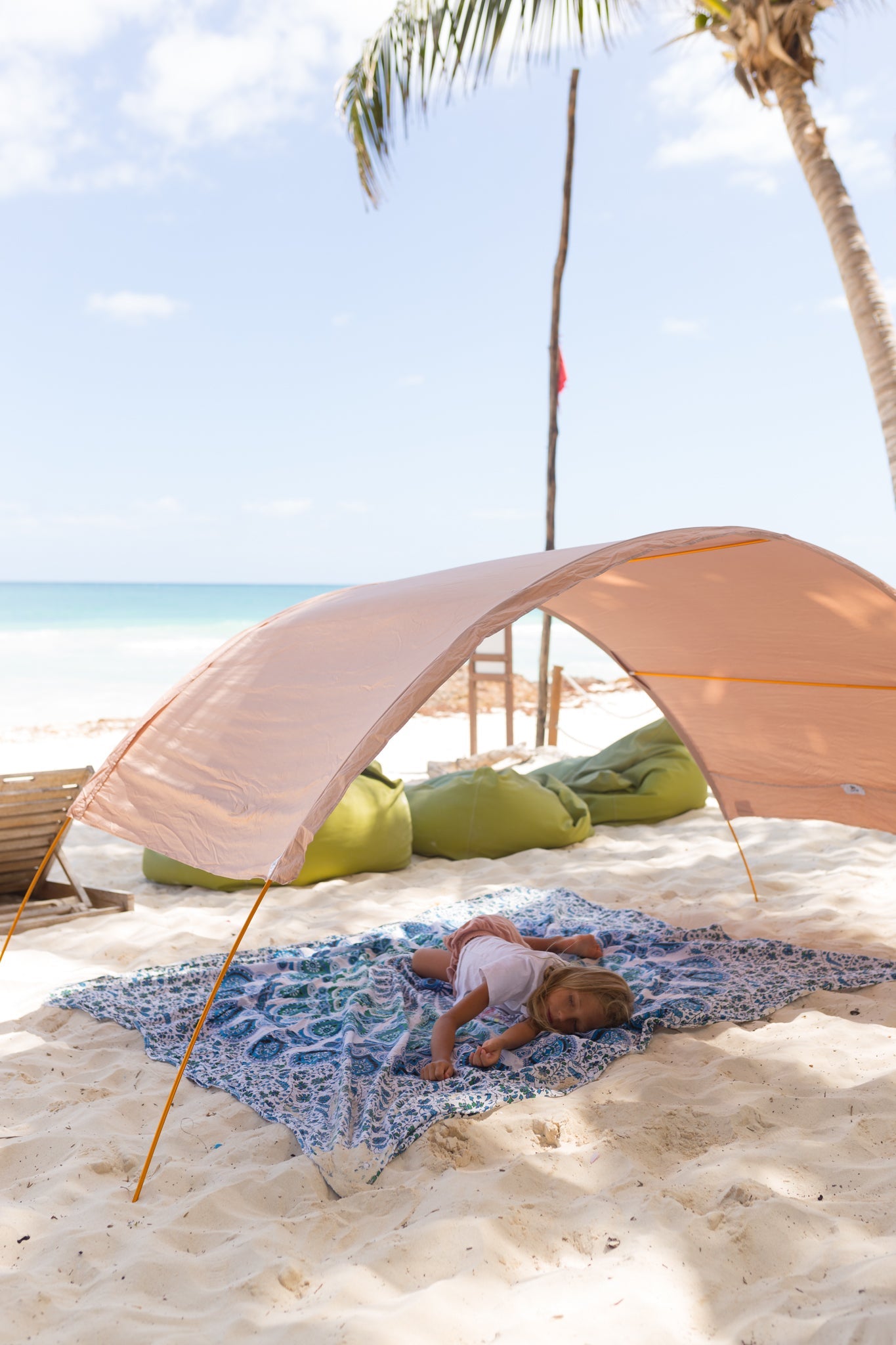 A child lies on a blue beach mat under a sand-coloured beach canopy on a sandy beach.
