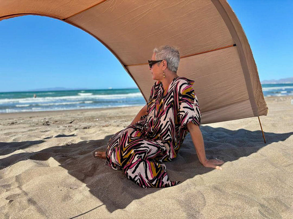 A woman with short gray hair sits on a sandy beach under a large beige beach cabana, wearing a loose, colorful abstract-patterned dress. The background shows the blue ocean and sky.