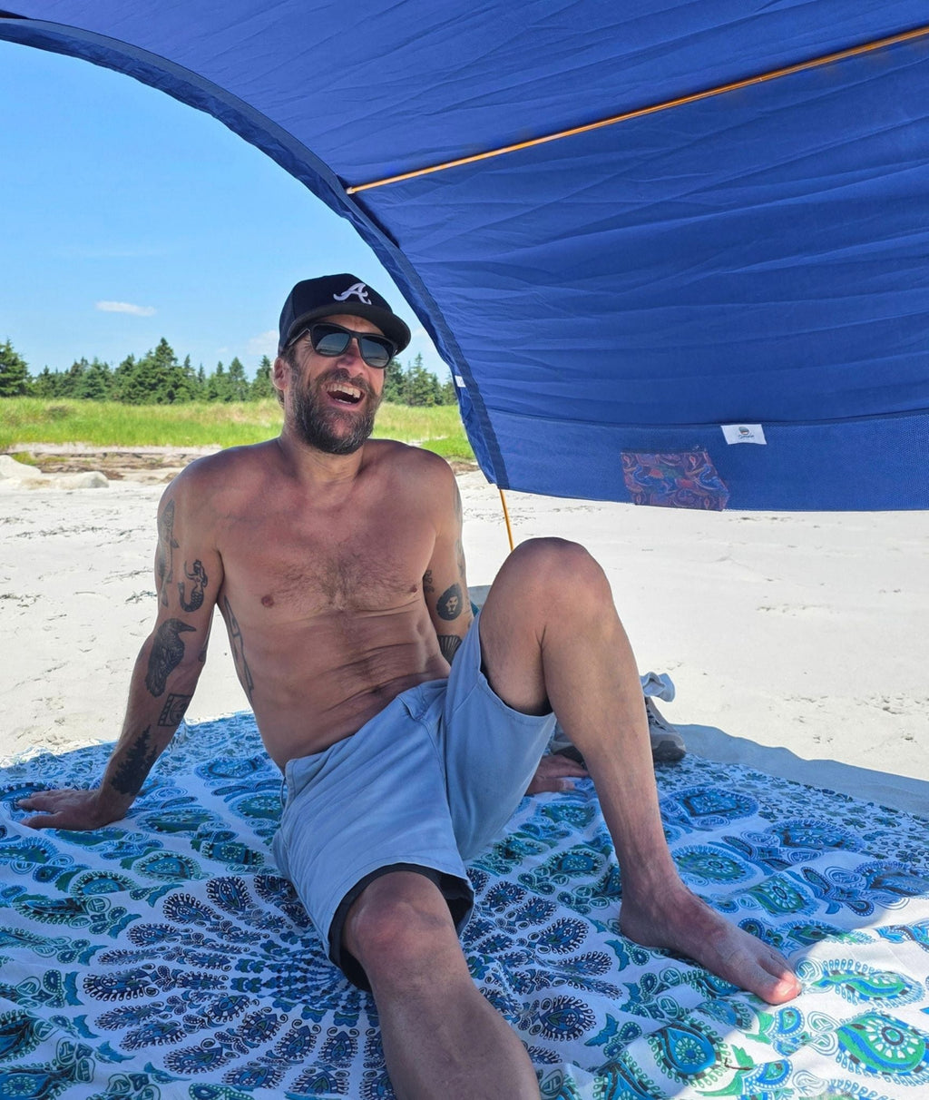 A man sitting shirtless on a mandala beach mat under a blue beach tent on a sandy beach.