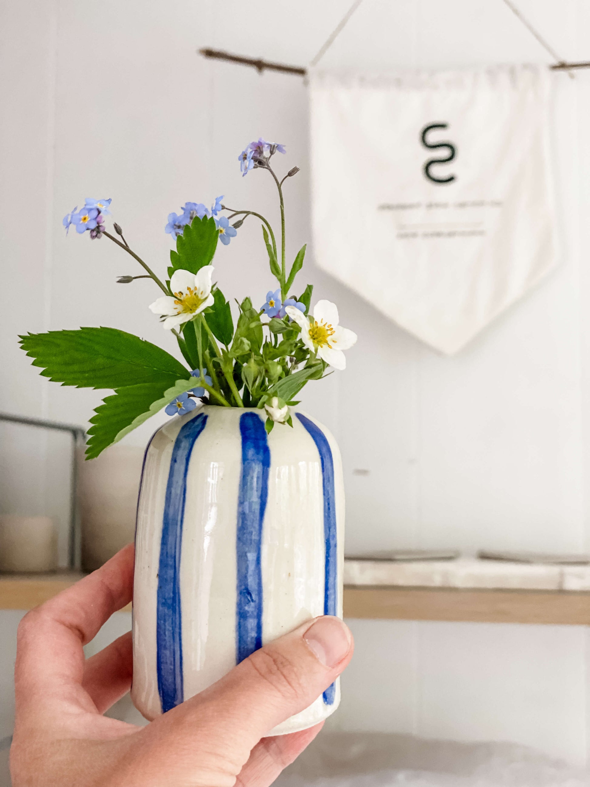 handmade pottery white porcelain bud vase hand painted blue stripes filled with dainty white, blue and yellow flowers, held in front of Steaped Studio Ceramics sign