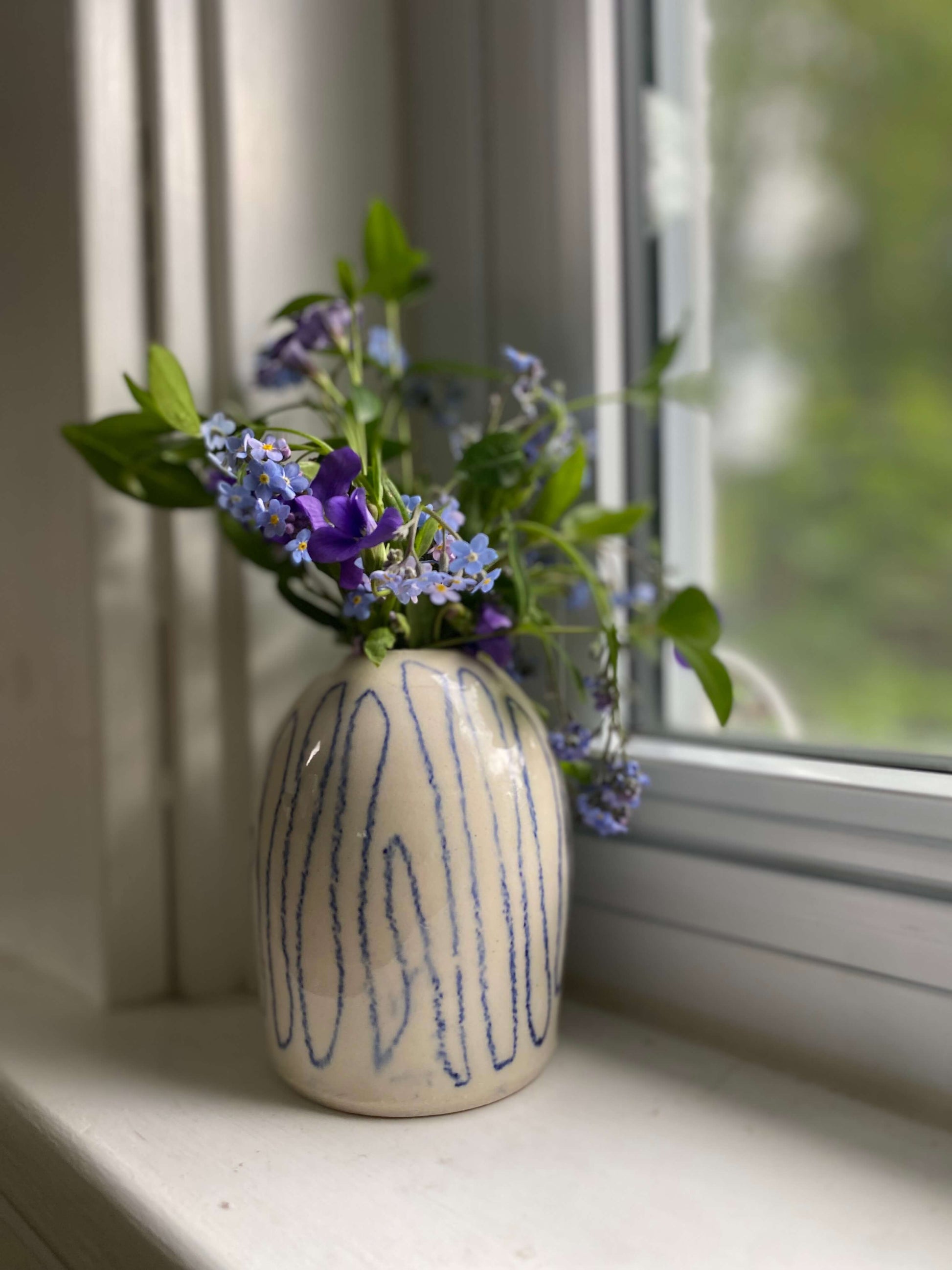 handmade pottery white porcelain bud vase hand drawn topographic line design filled with dainty blue and purple flowers, on window sil