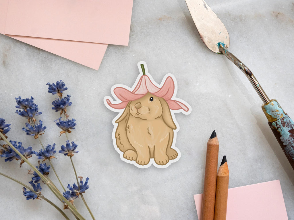 A sticker of an illustration of a bunny on a marble tile. The bunny is sitting facing forward with their head slightly turned to the side. The sticker is on a white marble tile and there's a palette knife and pencils to the right of it. To the left of the sticker, there are pink pieces of paper and dried lavender flowers.