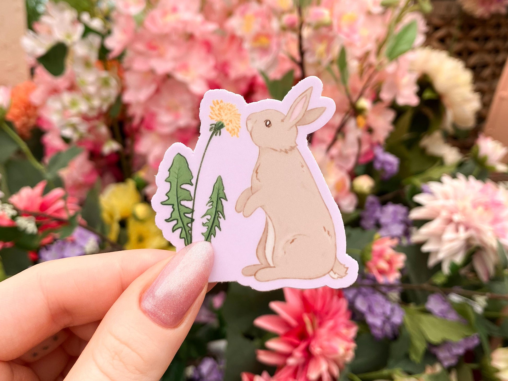 A white woman's hand holding a sticker of an illustration of a bunny in front of some flowers. The bunny is light brown and is standing on their hind legs reaching up to a dandelion flower. The background of the sticker is light purple.