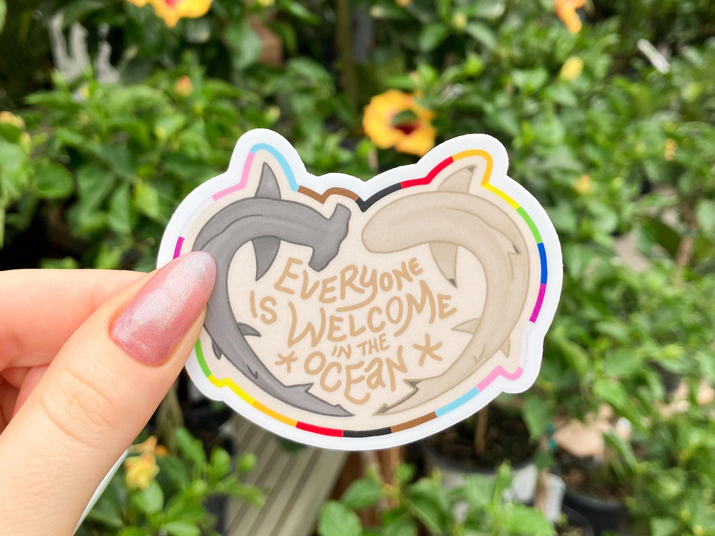 A white woman's hand holding a sticker with an illustration of two sharks in front of some flowers. One shark is a hammerhead shark and the other shark is a black-tip shark. The sharks form a heart. In the centre of the heart, there is text that says, "Everyone is welcome in the ocean". A rainbow border in the colours of the pride flag goes around the outside of the sticker.