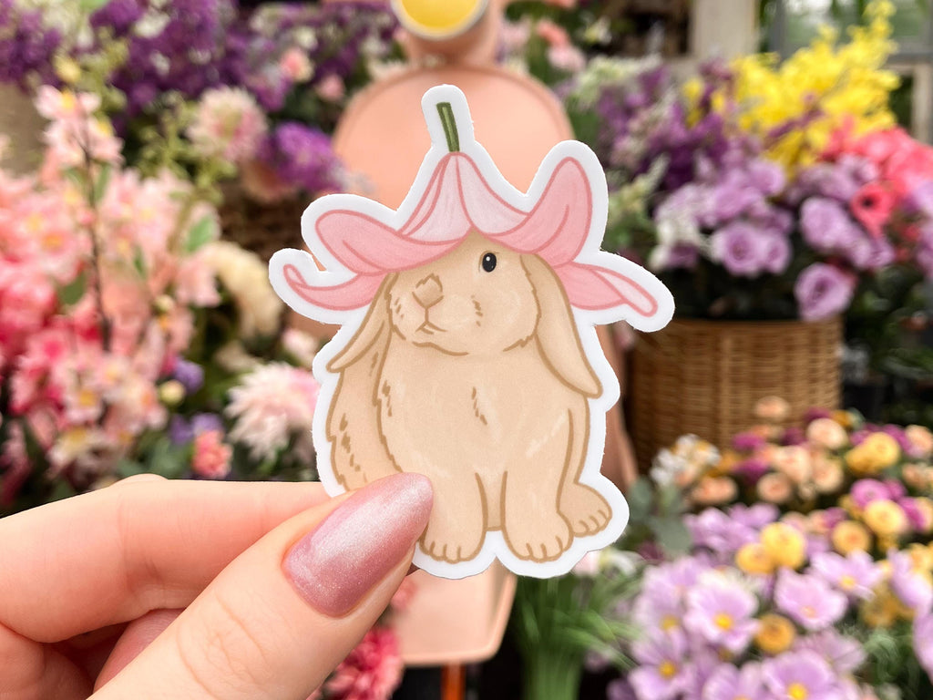 A white woman's hand holding a sticker of an illustration of a bunny in front of some flowers. The bunny is sitting facing forward with their head slightly turned to the side. They're wearing a pink flower hat.