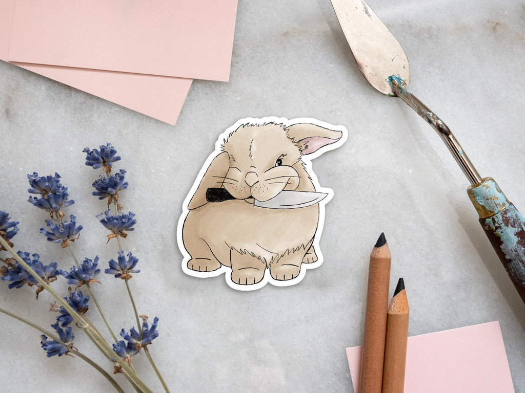 A sticker of an illustration of a light brown bunny holding a knife. The sticker is on a white marble tile and there's a palette knife and pencils to the right of it. To the left of the sticker, there are pink pieces of paper and dried lavender flowers.