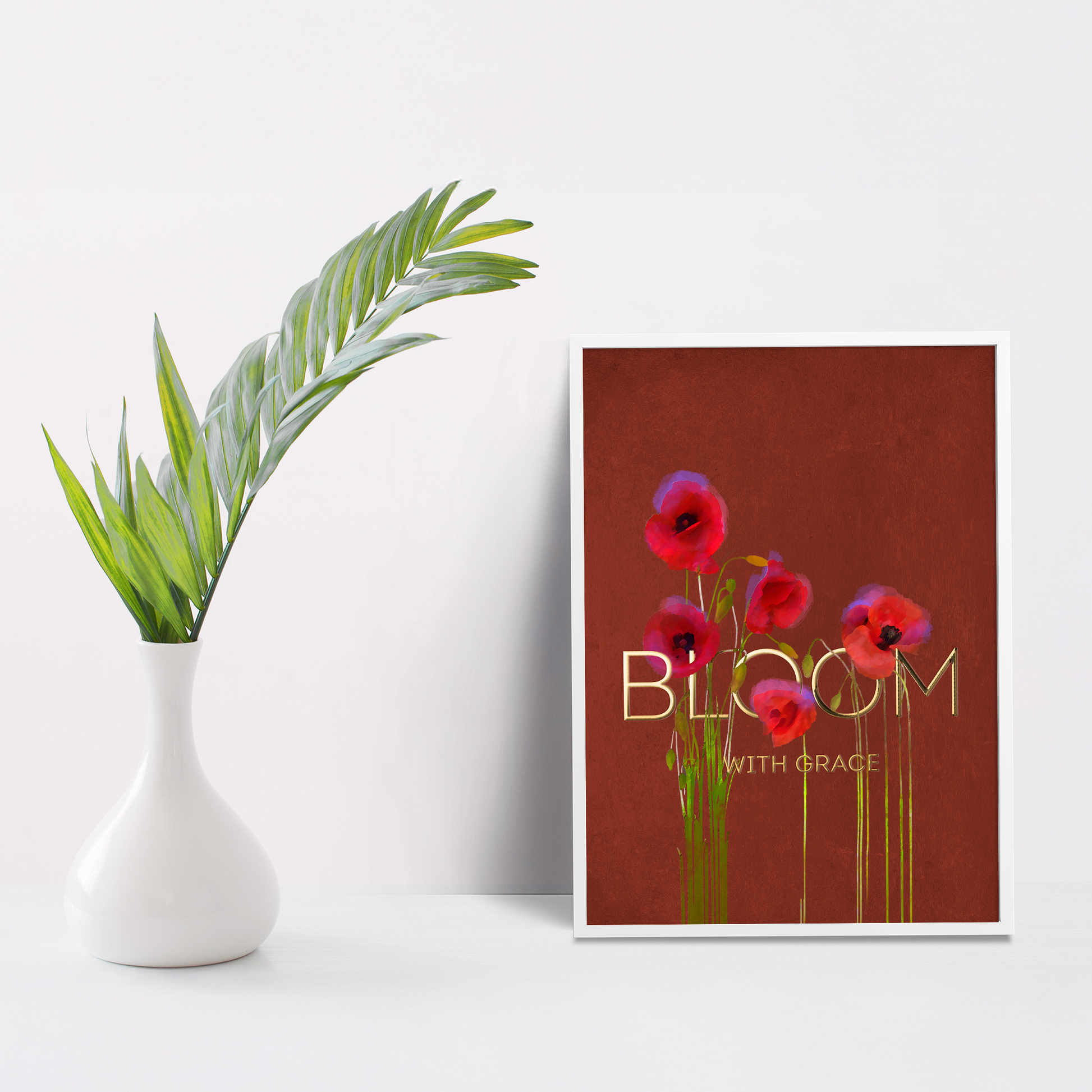 Framed artwork with red flowers and 'Bloom with Grace' text on a white surface with a vase and plant.