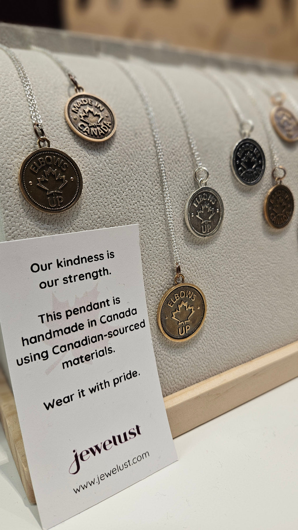 Many coin pendants, of different sizes, with 'Elbows Up' or 'Made in Canada' and a maple leaf design on a necklace display with a card describing the pendants