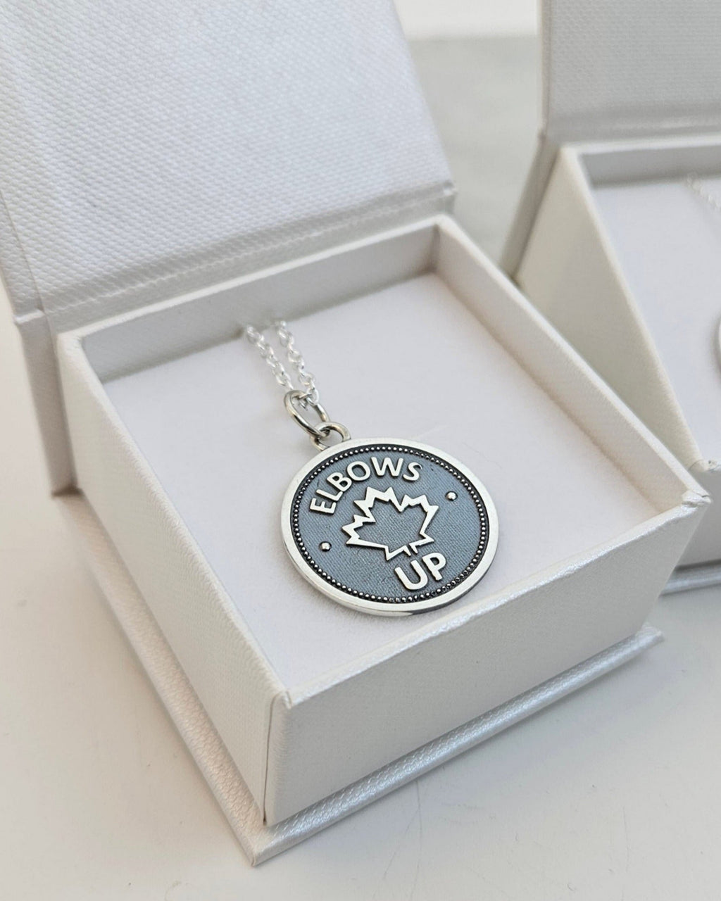 Silver coin pendant with 'Elbows Up' and a maple leaf design, on a silver chain, in a white jewellery box on a white background.