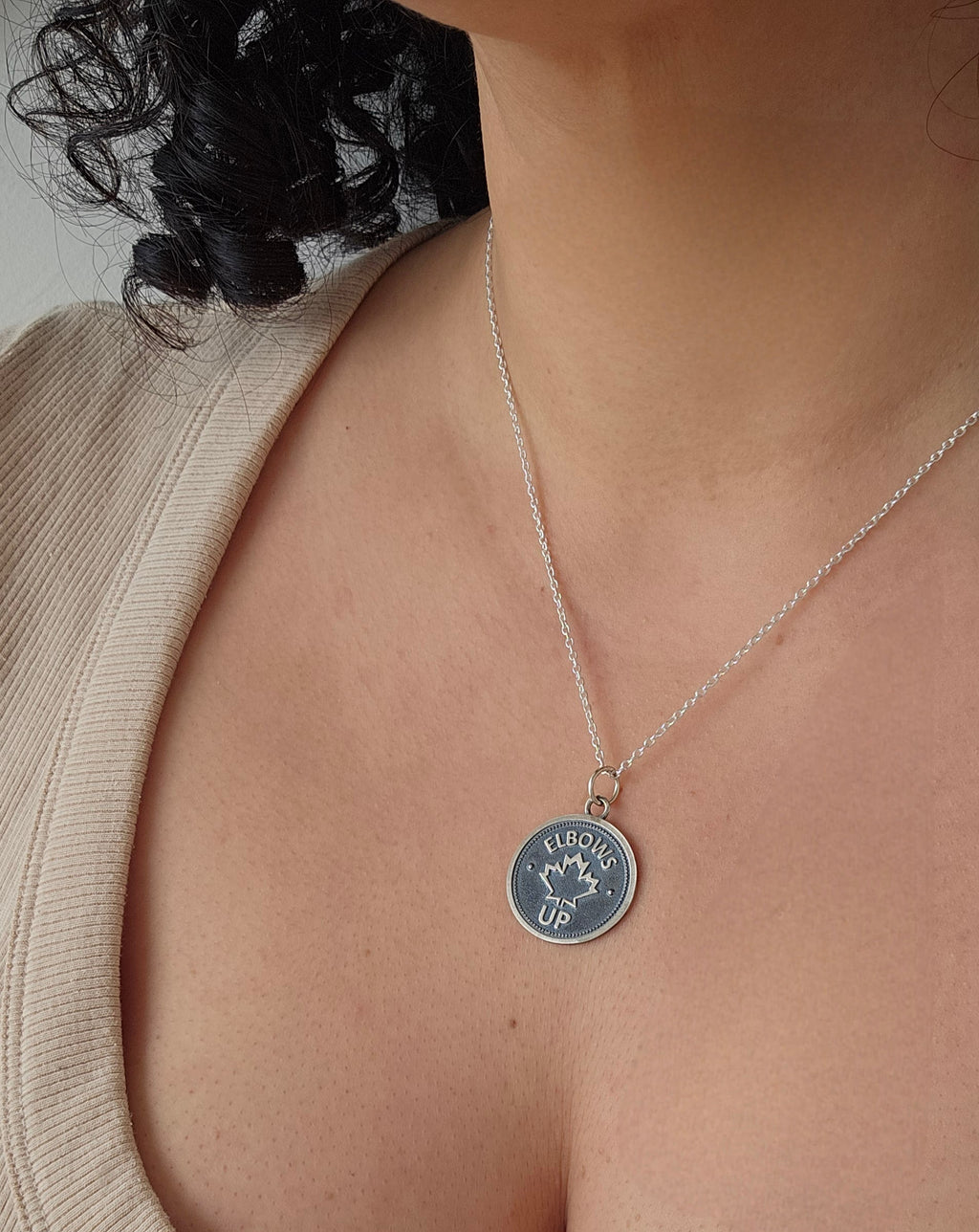 Coin pendant with 'Elbows Up' and a maple leaf design, on a silver chain, displayed on a model wearing a scoop-necked beige top.