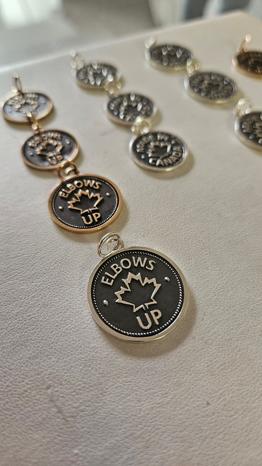 Many coin pendants, of different sizes and metals, with 'Elbows Up' and a maple leaf design, on a white surface