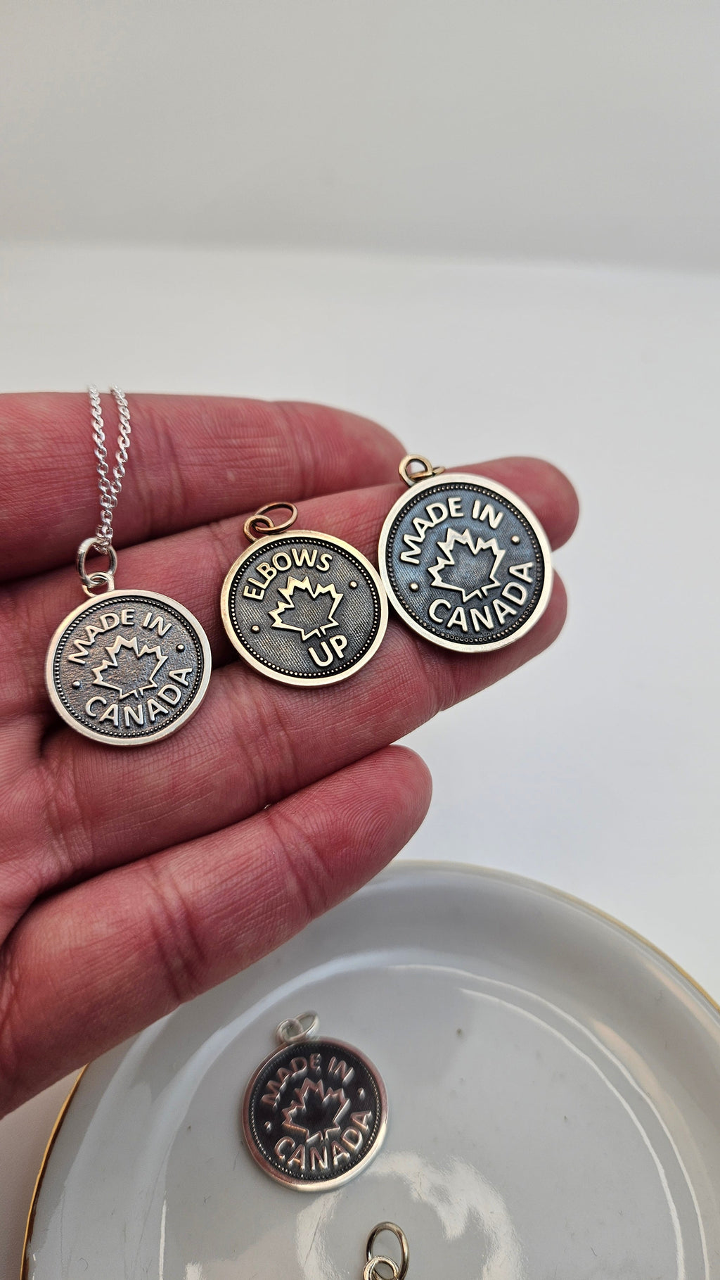 Three coin pendants, of different sizes, with 'Elbows Up' or 'Made in Canada' and a maple leaf design, in gold on the model's hand