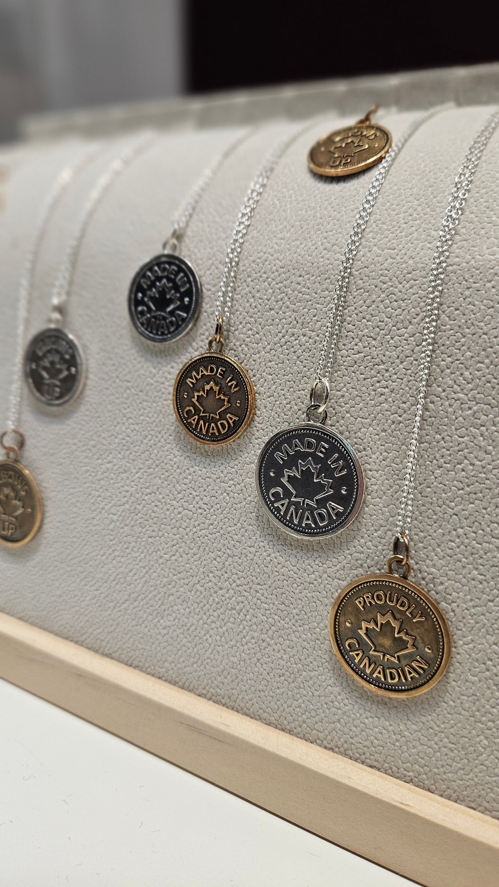 Many coin pendants, of different sizes and metals, with 'Elbows Up' or 'Made in Canada' or 'Proudly Canadian' and a maple leaf design on silver chains on a necklace display.